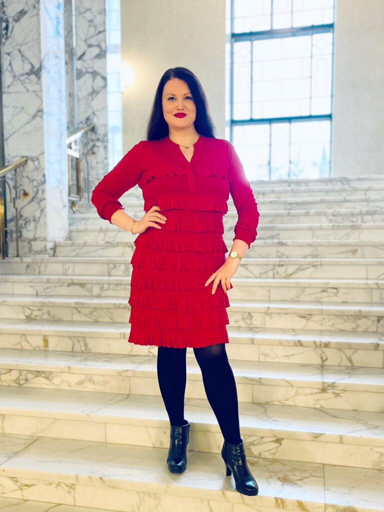 Tytti seisoo eduskuntatalon marmoriportaissa punaisessa mekossa, mustissa sukkahousuissa ja mustissa nilkkureissa hymyillen kameralle